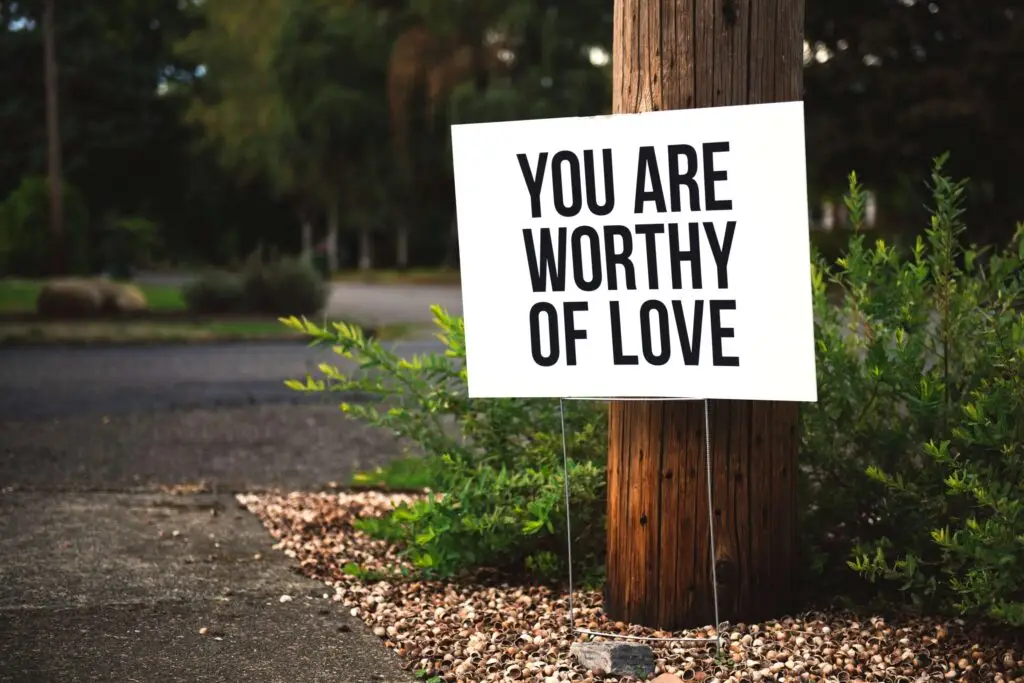 A white sign with bold black text reading YOU ARE WORTHY OF LOVE is attached to a wooden pole, serving as a gentle reminder in combatting suicide. It's surrounded by green shrubs and gravel, with a blurred street and trees in the background.