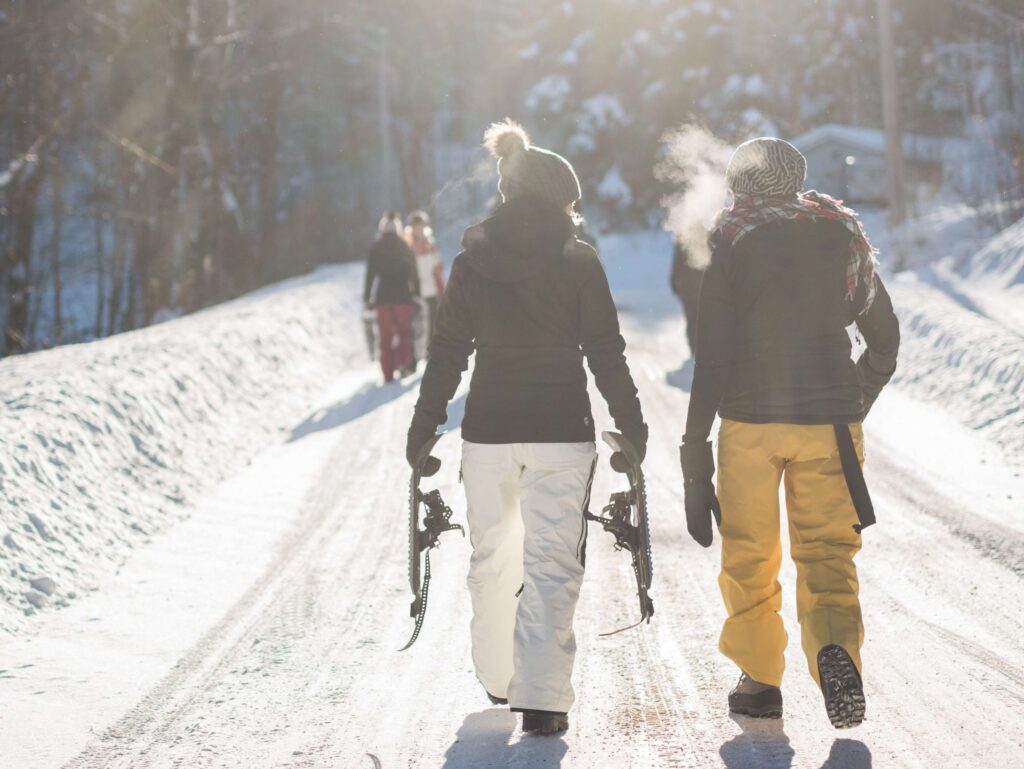 Two people in winter clothing traverse a snow-covered path, one carrying snowshoes. In the background, others enjoy winter games for kids under the bright sun, which casts a warm glow and makes breath vapor visible in the crisp air.