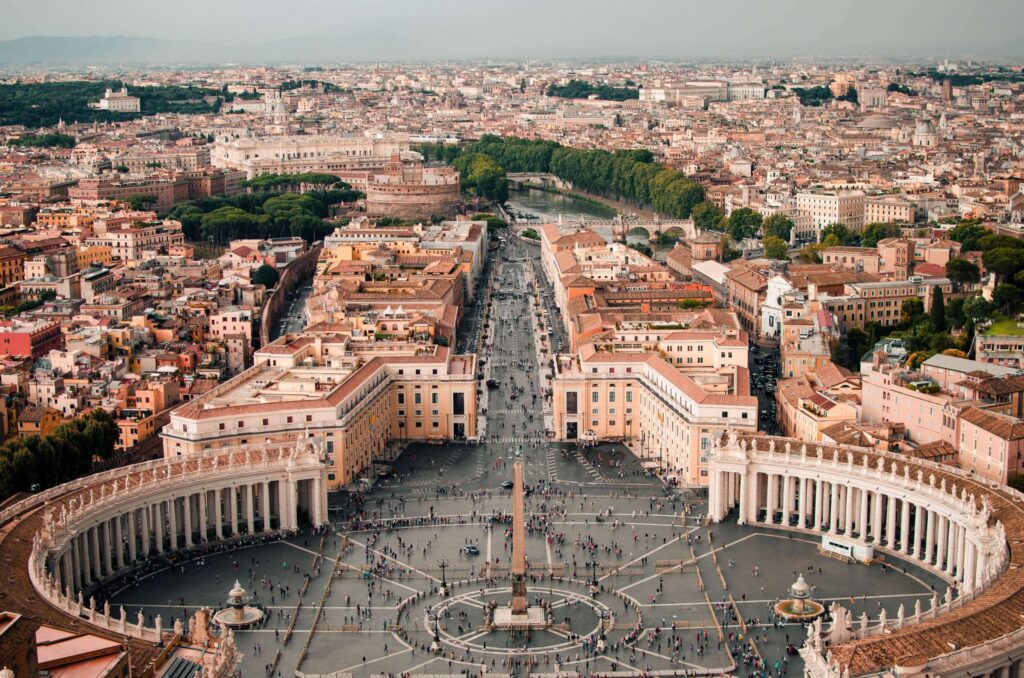 Aerial view of St. Peter's Square in Vatican City, with the iconic colonnades embracing the lively plaza. Dive into Rome travel tips as you marvel at the bustling crowd and gaze over Rome, where the Tiber River winds through its historic splendor.