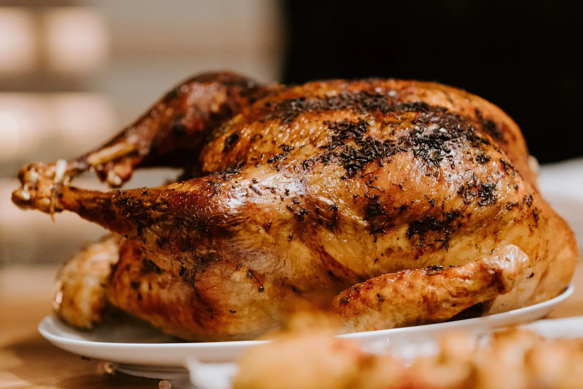 A roasted turkey on a white plate, seasoned with herbs and displaying golden, crispy skin, is ready to delight. Placed on a wooden surface, it sets the scene for a warm and inviting Thanksgiving meal that inspires laughter like riddles for kids around the table.