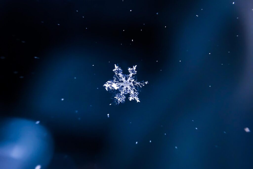 Close-up of a single snowflake against a dark blue background, perfect for winter trivia for kids. The snowflake is intricately detailed with symmetrical patterns, surrounded by small white specks resembling falling snow.