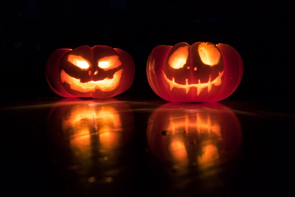 Two glowing jack-o'-lanterns with carved faces sit side by side in the dark, embodying the very essence of Halloween. Their light casts reflections on a shiny surface below, creating an eerie yet festive atmosphere.