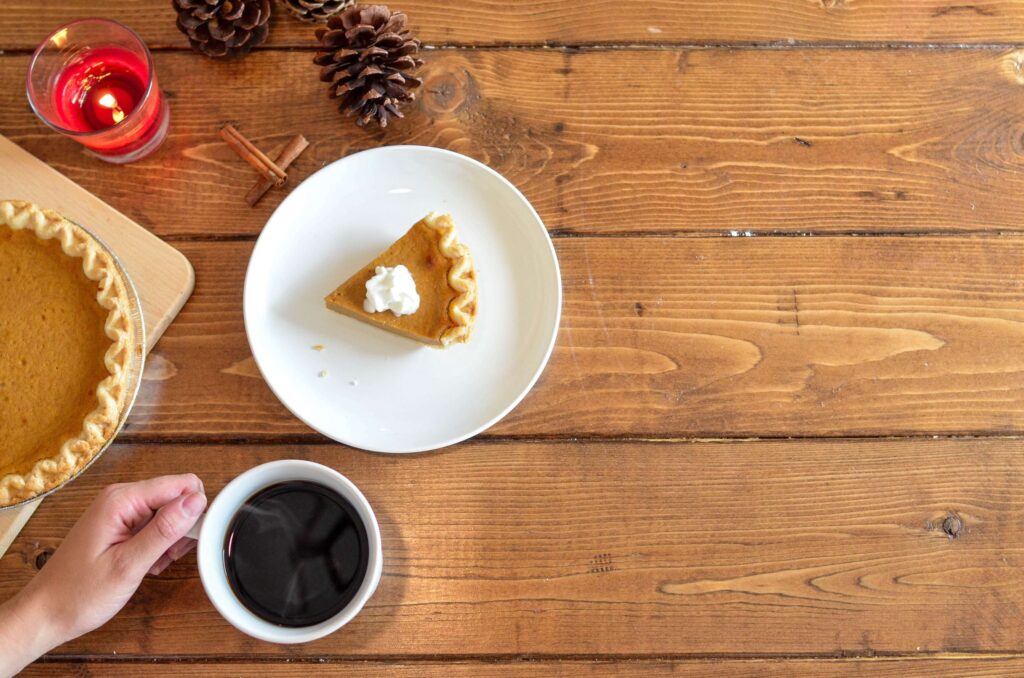 A hand holding a cup of coffee sits next to a slice of pumpkin pie topped with whipped cream on a plate. A whole pie, pinecones, and a red candle create an inviting autumn setting on the wooden table, perfect for sharing Thanksgiving moments and considering gifts for kids.