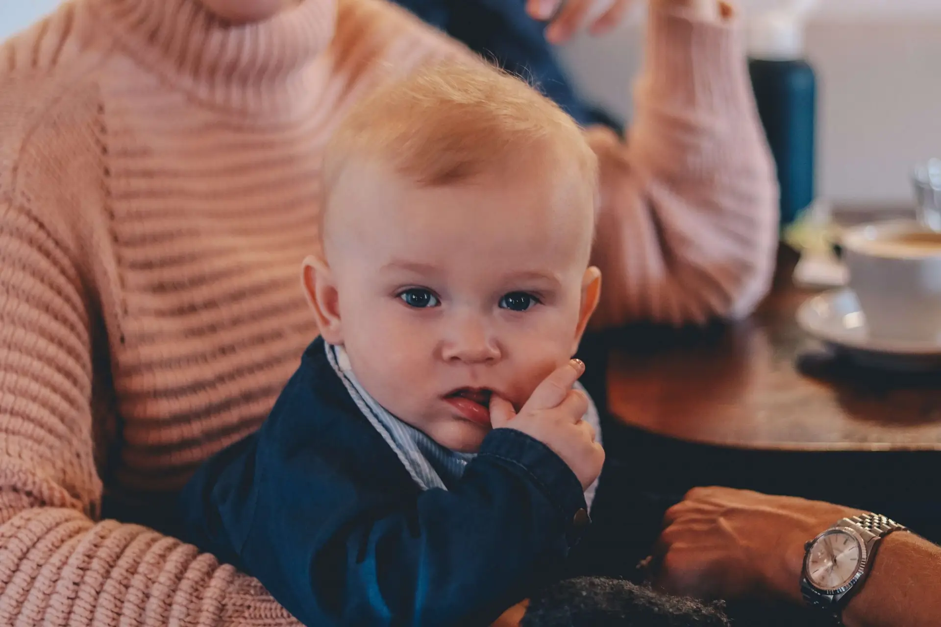 A baby with light hair, perhaps pondering why kids suck their thumb, sits on an adult's lap in a dark blue outfit, sucking on their finger. The adult in a pink knit sweater enjoys coffee while checking their smartwatch at the table beside them.