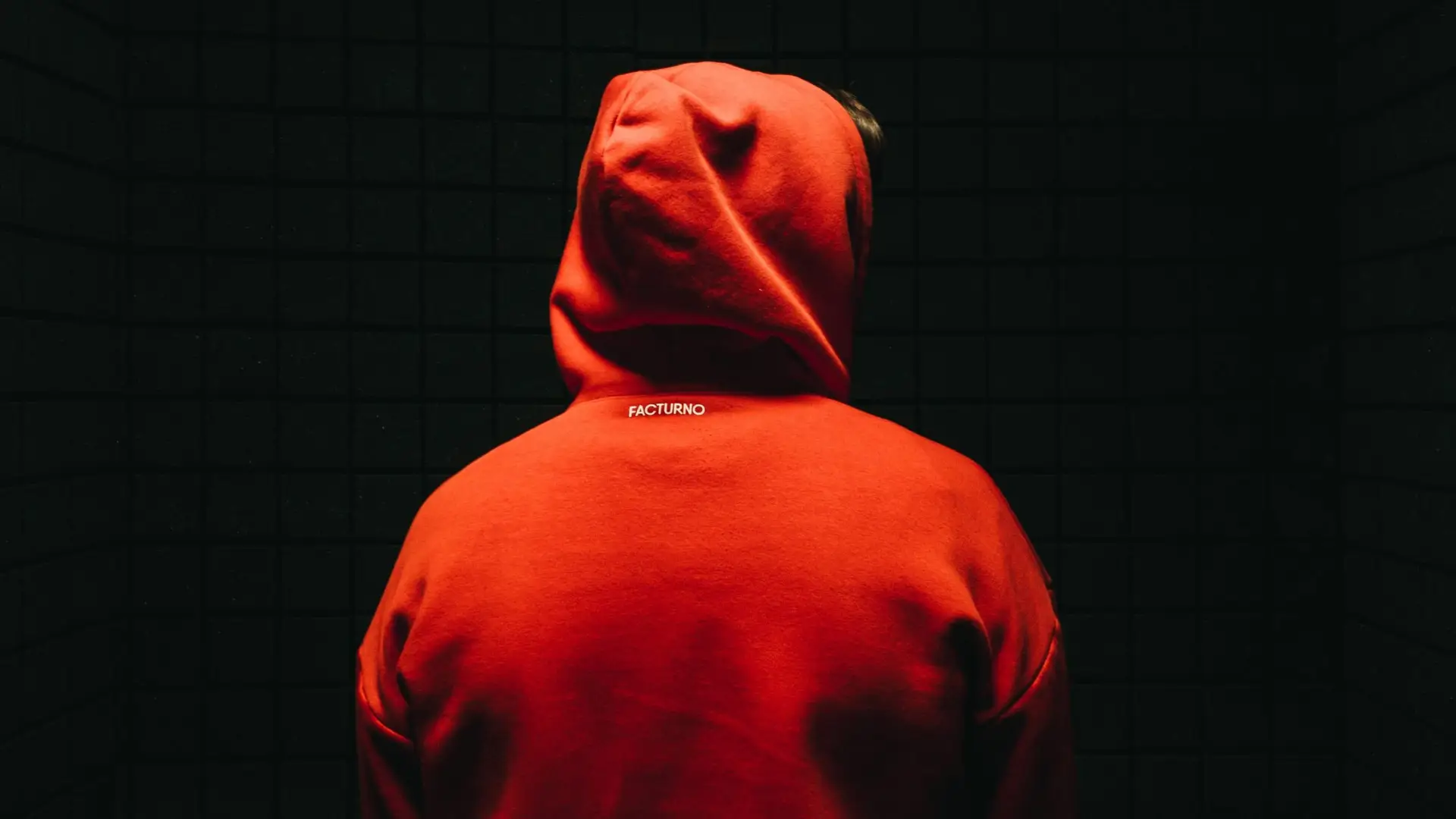 A person in a bright red hoodie stands facing a dark, tiled wall. The hood is up, obscuring their face. The word PICTURNO is visible on the back of the hoodie, contrasting with the dimly lit background—a reminder of why teens wear hoodies even in the summer's heat.