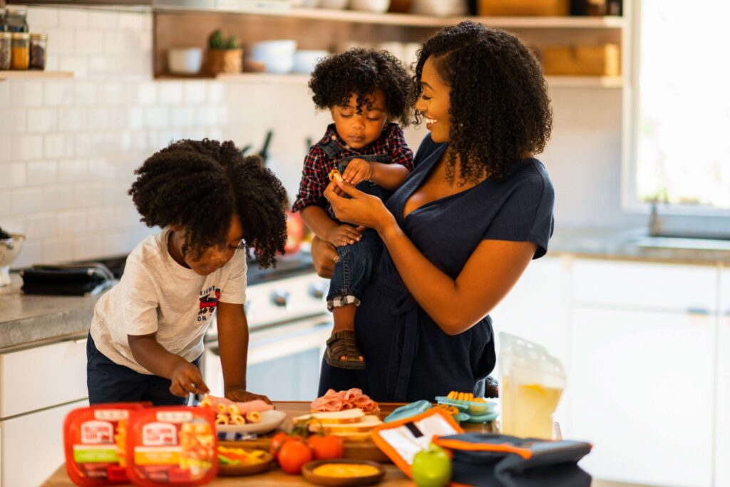 In a bright kitchen, a woman with two children prepares a meal, perhaps exploring kid-friendly Thanksgiving recipes. One child stands on a chair cutting food, while the other is held by the woman. The table is filled with ingredients and kitchen items. Everyone appears content and focused.