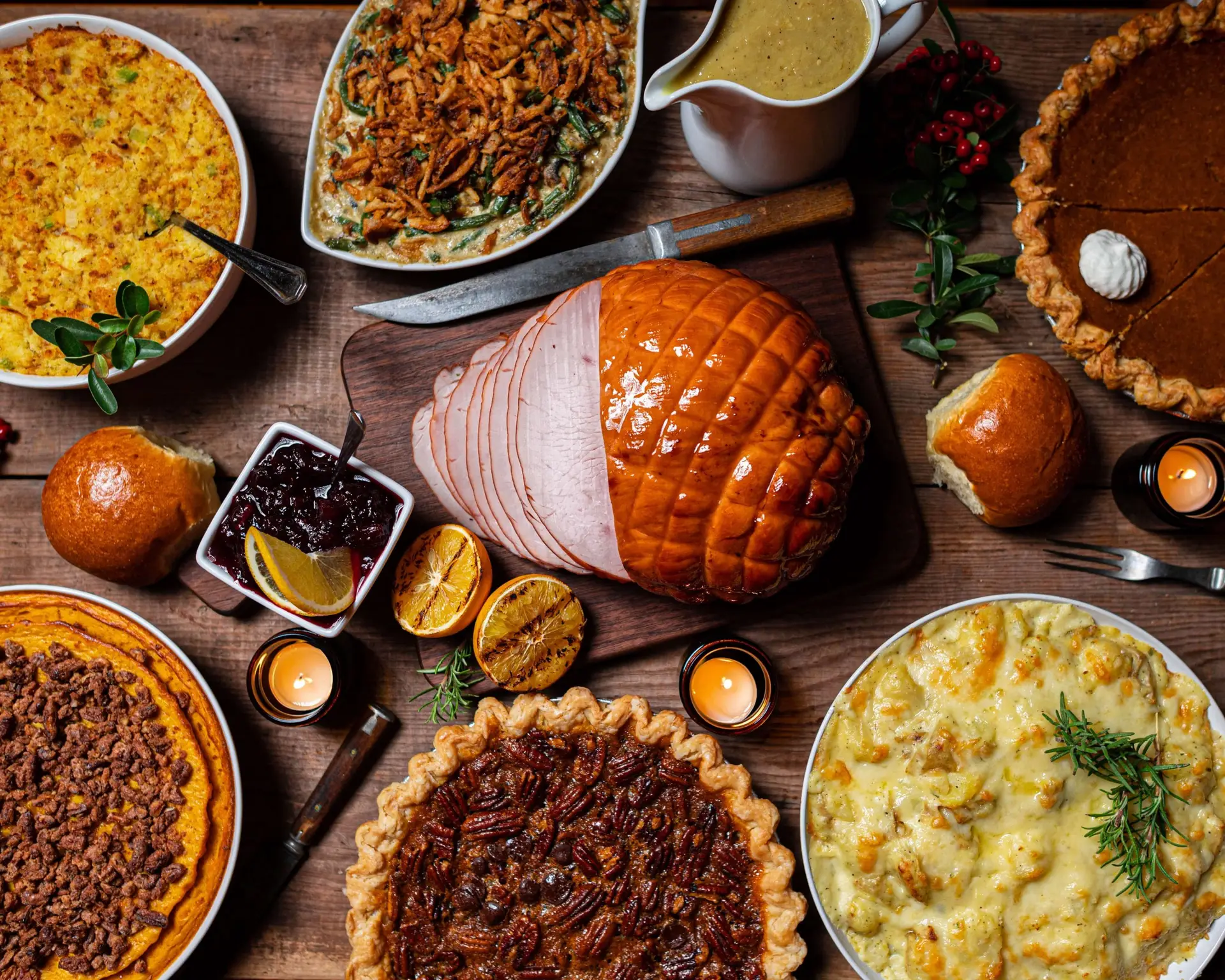 A festive table spread with a glazed ham at the center, surrounded by kid-friendly Thanksgiving sides like pies, mashed potatoes, and green bean casserole. Cornbread, cranberry sauce, bread rolls, and gravy round out the meal. Candles and festive decor complete the setting.