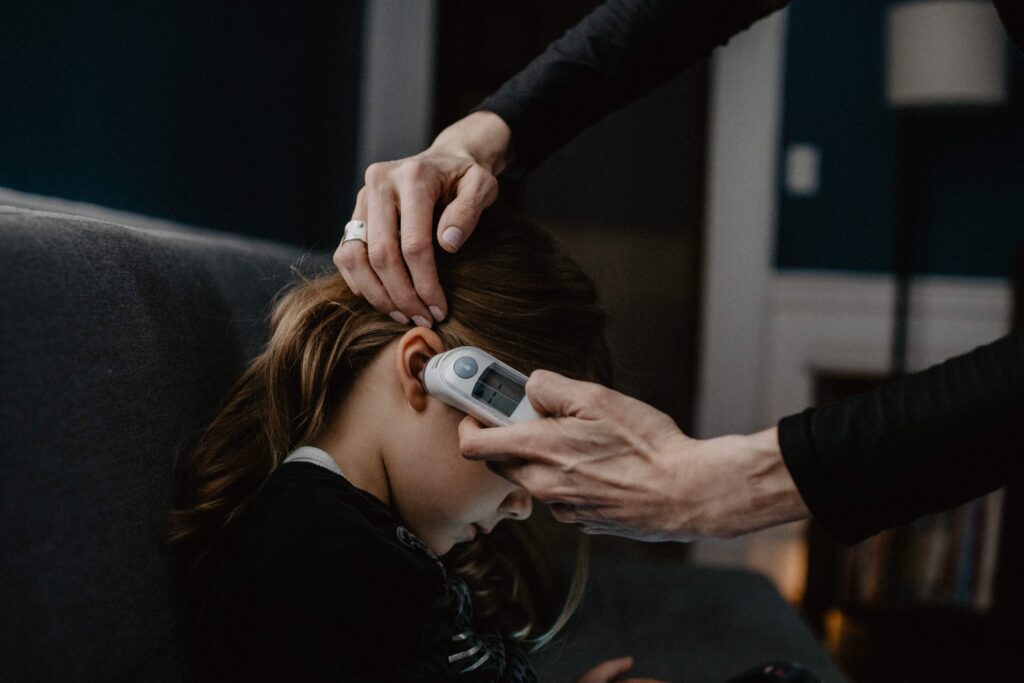 An adult's hands gently take the temperature of a young girl using an ear thermometer. As she sits quietly on a sofa, looking down, the dim lighting creates a cozy atmosphere, inviting thoughts about why kids get sick so often in such serene moments.