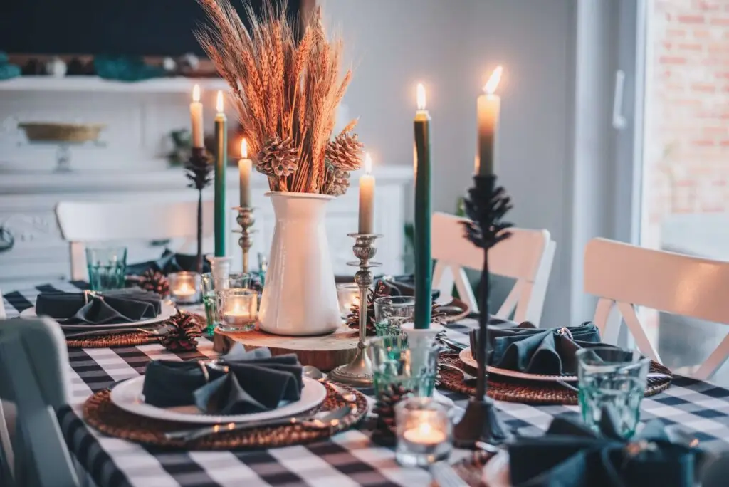 A dining table is elegantly set with a black and white checkered tablecloth, dark napkins, and candlelit centerpieces in pine cone holders. A white vase with dried plants sits in the center, creating a cozy atmosphere perfect for sharing Thanksgiving jokes for kids.