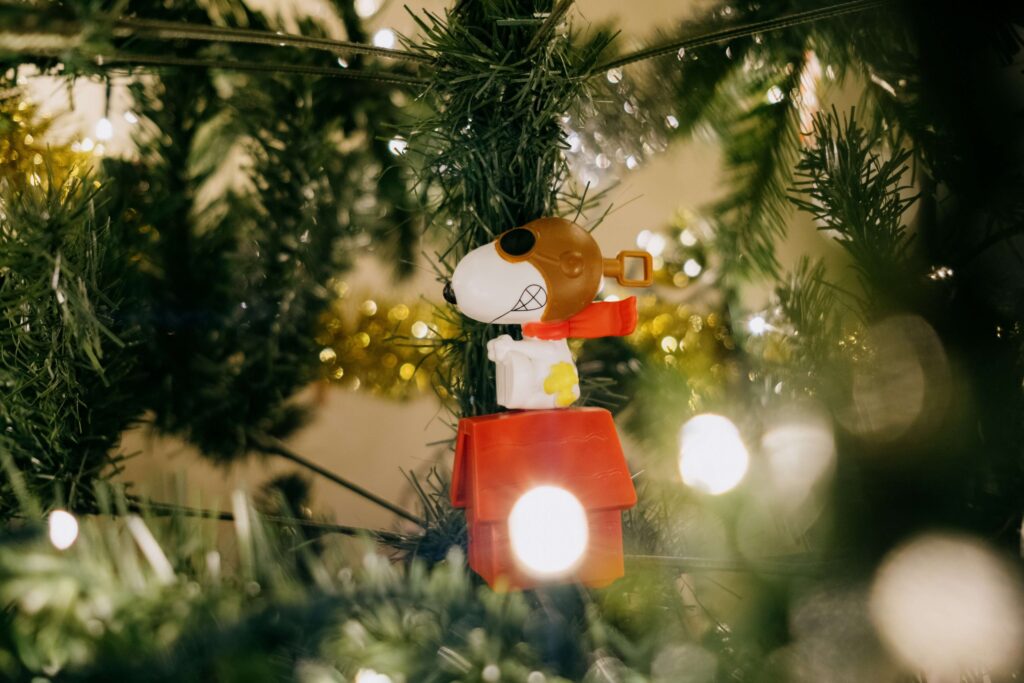 A figurine of a beagle wearing a red scarf and goggles is perched on top of a small red doghouse, reminiscent of classic Thanksgiving kids movies, nestled among green Christmas tree branches and glowing white lights.