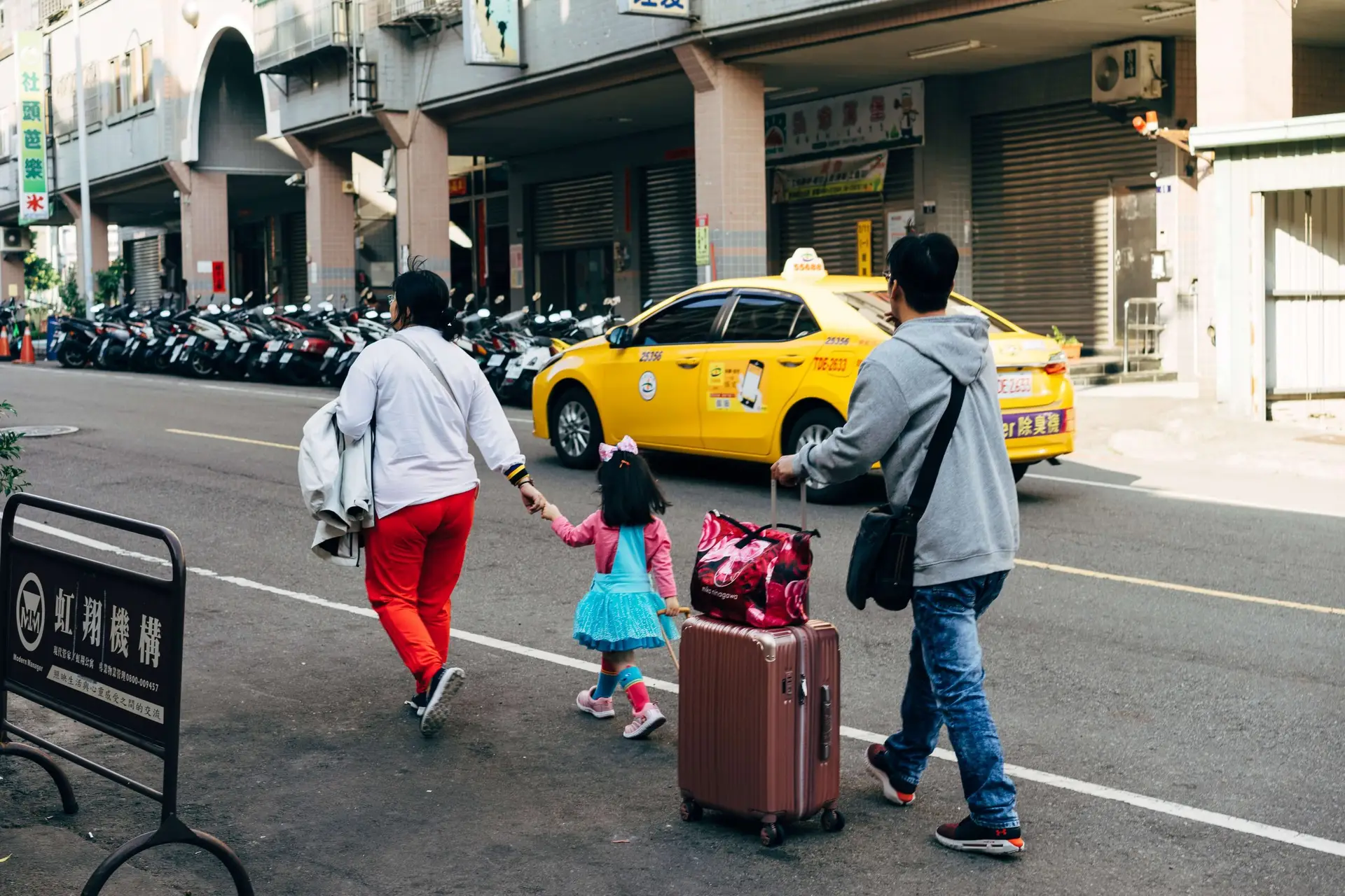 A family strolls down a city street, with a man pulling a suitcase and holding a child's hand, showcasing practical kids travel accessories. A woman carries a backpack while parked scooters and a yellow taxi line the scene. The child wears a blue dress and pink backpack.