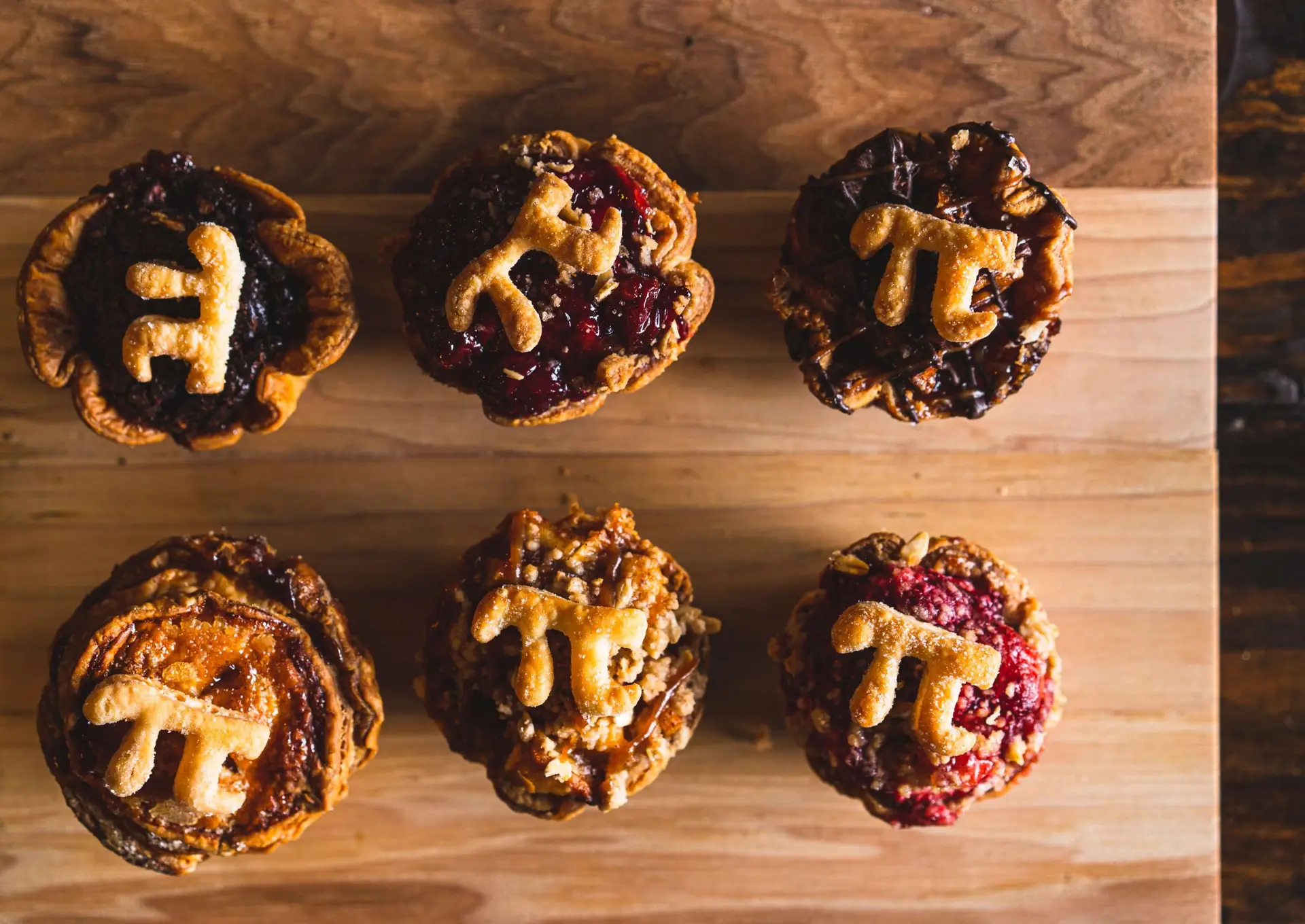 Six delicious mini pies rest on a wooden surface, each crowned with a golden crust shaped like the Pi symbol (π). Perfect thanksgiving desserts for kids, these pies boast various fillings like berry and pecan, offering a delightful blend of colors and textures.