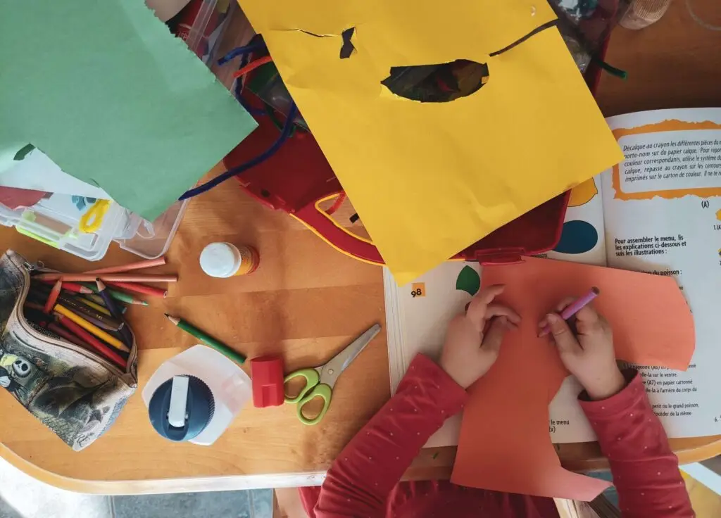 A child in a red shirt cuts orange construction paper at a table brimming with art supplies, from colored pencils to a curious open book. Perhaps they ponder why kids sometimes eat paper amid the vibrant chaos of creativity.