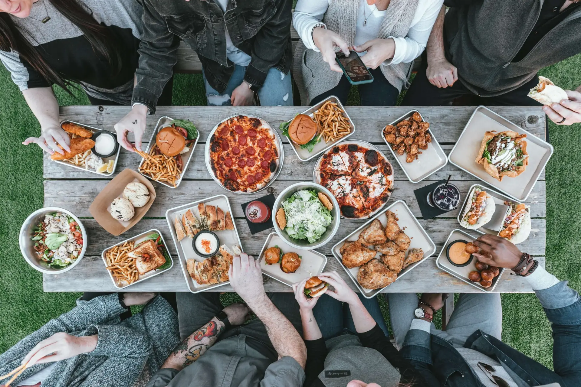 A group of people sitting around a wooden table filled with various foods, including pizza, burgers, fries, salads, chicken wings, and drinks. Like a feast of Thanksgiving treats for kids, they are eagerly reaching for food and joyfully interacting with each other.