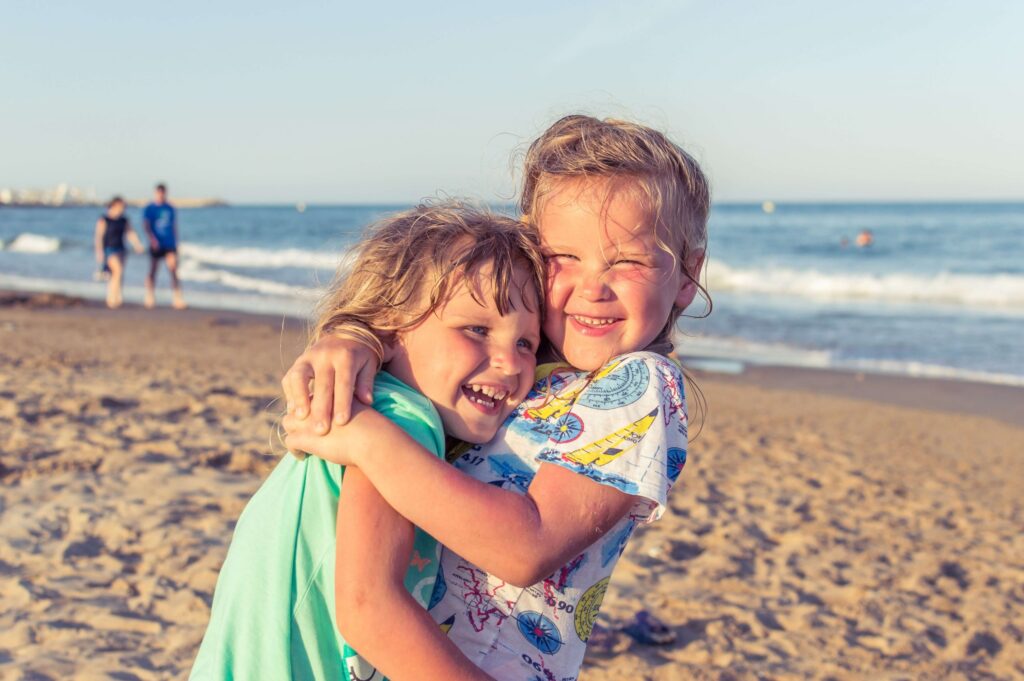 Two young children, engaged in joyful travel activities for kids, happily embrace on a sandy beach. The child on the left wears a turquoise shirt, while the one on the right sports a colorful shirt with a map pattern. The ocean and a few people are in the background under a clear sky.