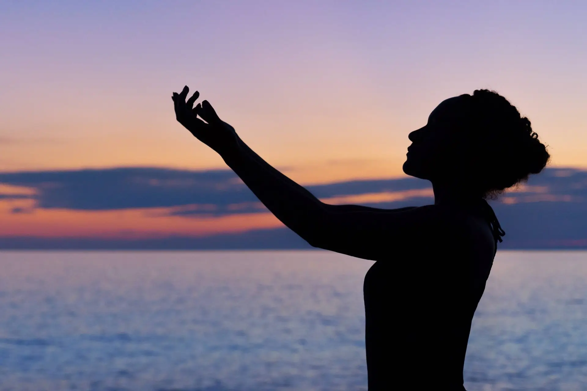 Silhouette of a person with curly hair raising their hands towards the sky, celebrating World Mental Health Day by the sea during a colorful sunset, with hues of orange, purple, and blue.