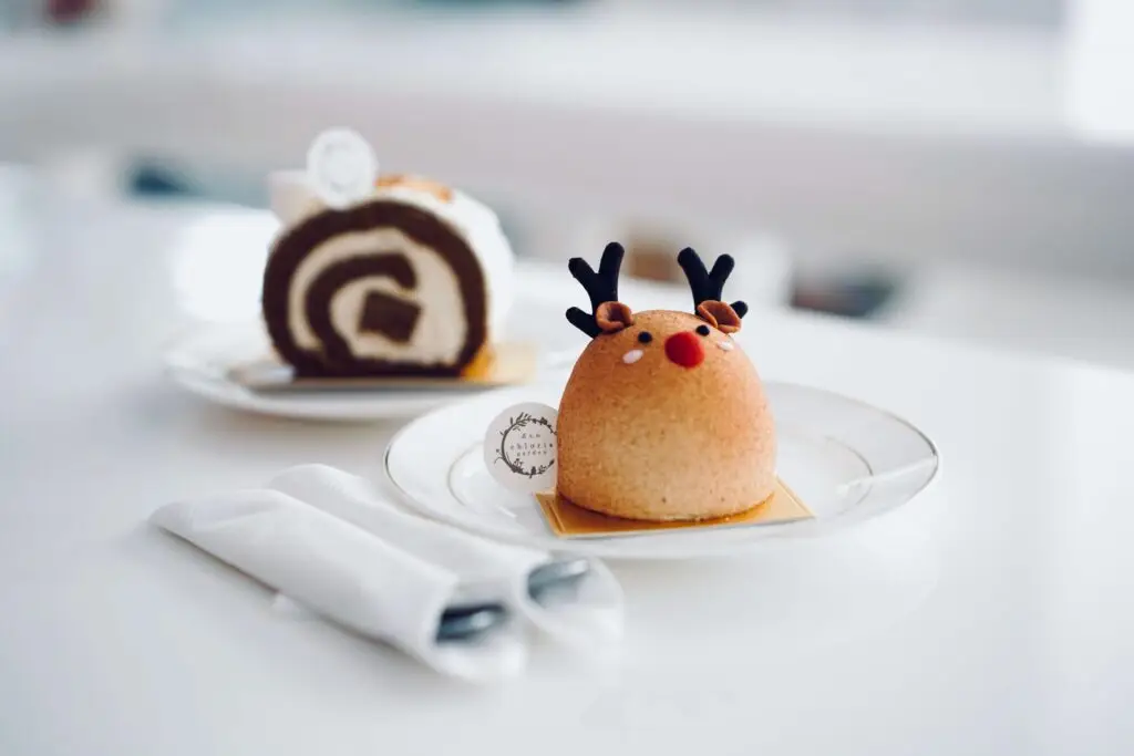 A round, reindeer-themed Christmas dessert on a white plate, featuring chocolate antlers and a red nose, sits in the foreground. In the background, a slice of chocolate swirl cake is on another plate. Two rolled napkins with cutlery are beside the plates.