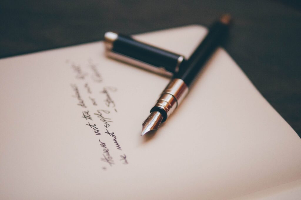 A fountain pen with a silver nib rests on an open notebook filled with the beginnings of winter poems for kids. The pen's cap is removed and lies beside it. The paper shows partly visible text, as the softly blurred background creates a warm and cozy atmosphere.