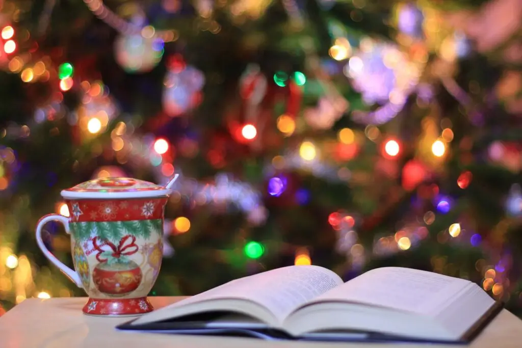 A festive mug with a lid and spoon sits next to a classic Christmas book on the table. In the background, colorful Christmas lights and decorations create a warm, blurred ambiance.