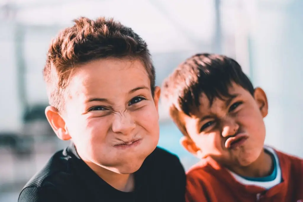 Two young boys making funny faces, perhaps inspired by some Thanksgiving jokes for kids; one with puffed cheeks and the other with a pout. They seem to be enjoying their playful moment outdoors. The background is softly blurred, highlighting the boys' expressions.