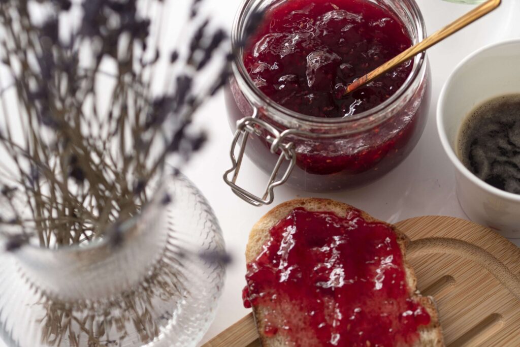 A jar of Christmas jam with a spoon inside sits next to a slice of bread spread with the festive treat on a wooden board. A cup of black coffee and a vase with dried lavender are nearby on the white surface.