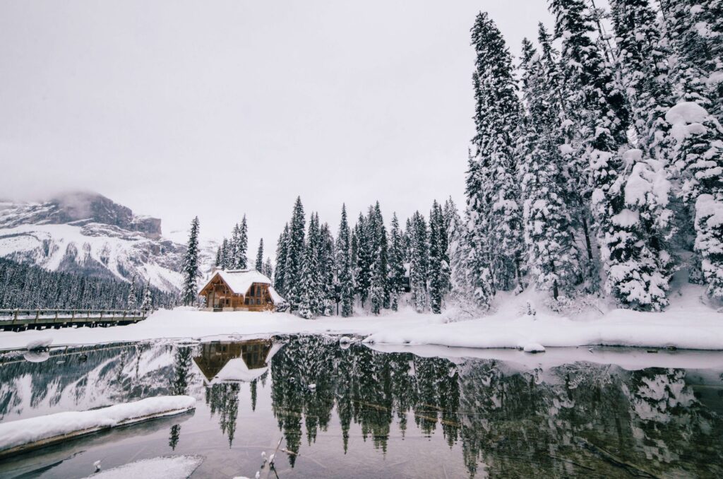 A snowy landscape with a wooden cabin serves as a perfect winter camp for kids beside a calm, reflective lake surrounded by tall, snow-covered pine trees. The overcast sky enhances the serene winter atmosphere.