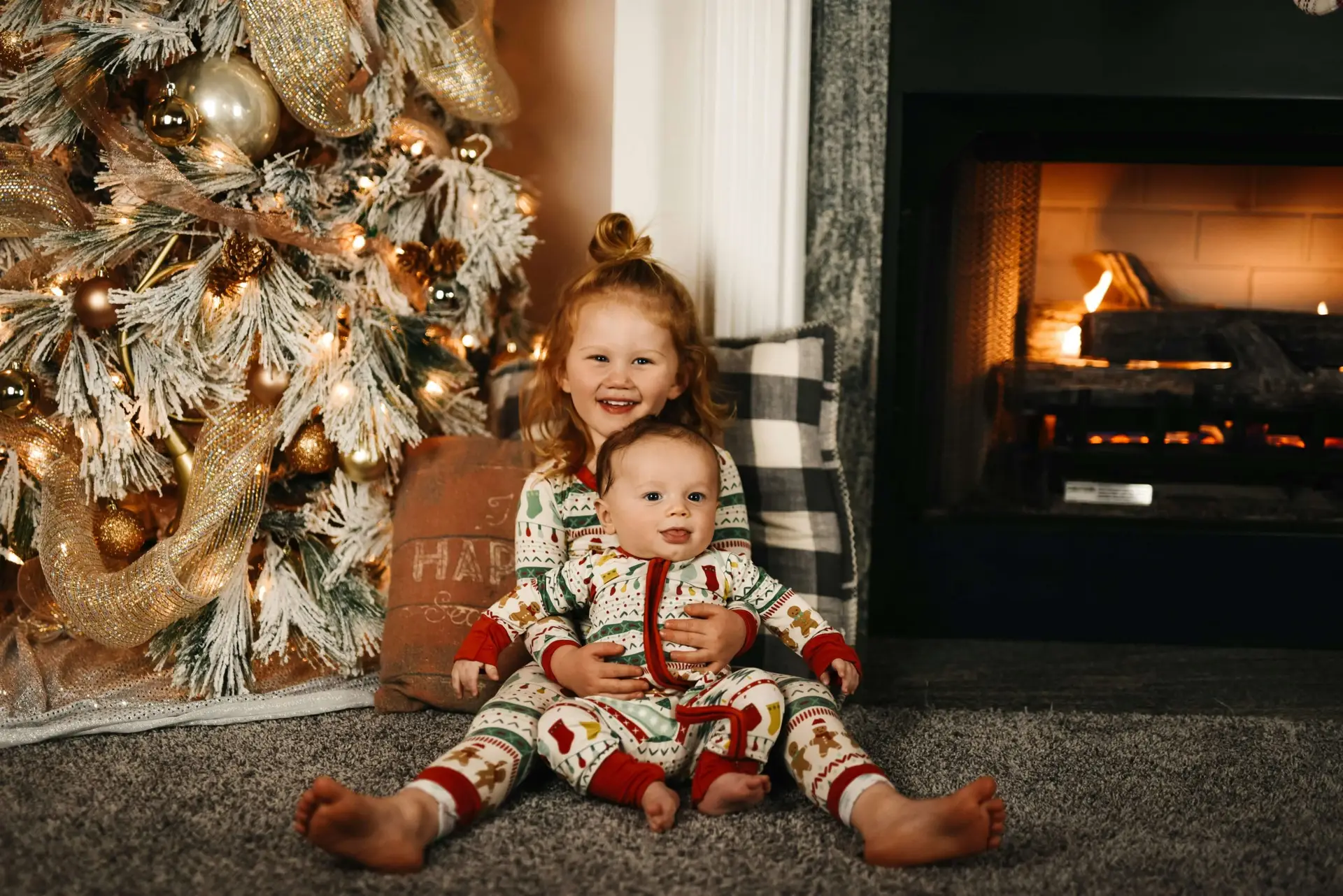 A young child with red hair sits on the floor in front of a lit fireplace, cradling a baby. Both don festive Christmas pajamas for kids adorned with holiday patterns. A decorated Christmas tree with lights and golden ornaments stands to the left. They are smiling joyfully.