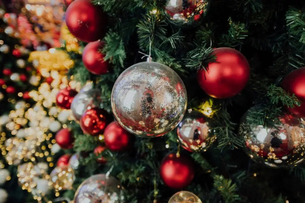 Close-up of a Christmas tree adorned with painted red and silver ornaments. The tree is lush with greenery, and warm white lights add a festive glow. The reflective silver baubles capture some surrounding lights and decorations.