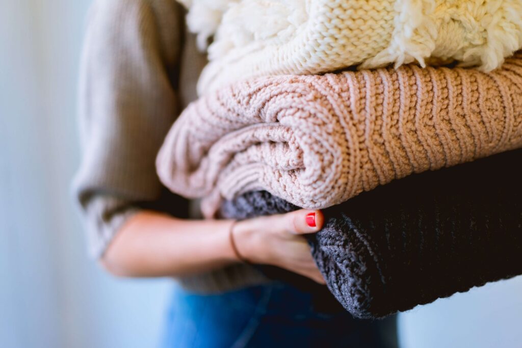 A person holding a stack of folded knitted blankets in cream, beige, and dark gray, perfect for cozy winter nights. The person is wearing a gray sweater reminiscent of Christmas sweaters for kids and has red nail polish. The background is blurred, adding to the warm ambiance.