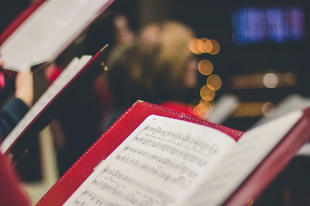 Close-up of red folders containing sheet music for winter songs for kids, held by choir members. The background is softly blurred with warm lighting, creating a cozy atmosphere.