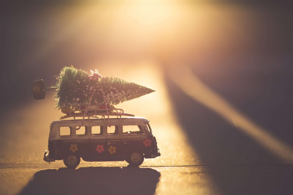 A small toy van with colorful flower patterns carries a miniature Christmas tree on its roof. The scene, set on a sunlit surface, twinkles with holiday cheer. It's a "tree-mendous" ride that's sprucing up the golden light, creating a pun-filled festive atmosphere!