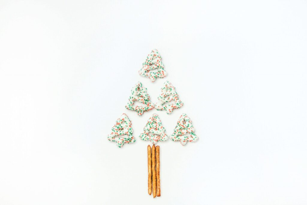 A delightful Christmas tree shape made of six frosted cookies with green and red sprinkles, arranged in a triangle on a white background. Perfect for christmas party snacks at school, pretzel sticks form the tree trunk at the bottom.