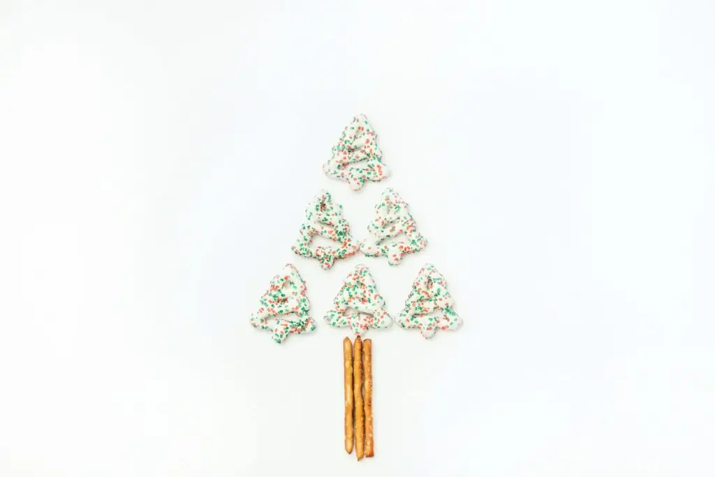 A delightful Christmas tree shape made of six frosted cookies with green and red sprinkles, arranged in a triangle on a white background. Perfect for christmas party snacks at school, pretzel sticks form the tree trunk at the bottom.