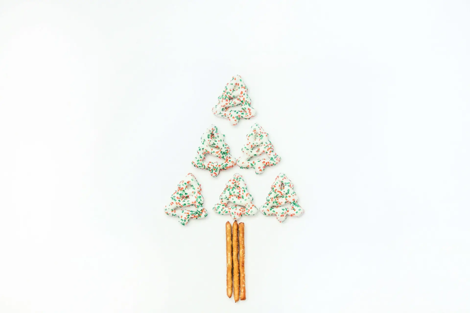 A delightful Christmas tree shape made of six frosted cookies with green and red sprinkles, arranged in a triangle on a white background. Perfect for christmas party snacks at school, pretzel sticks form the tree trunk at the bottom.