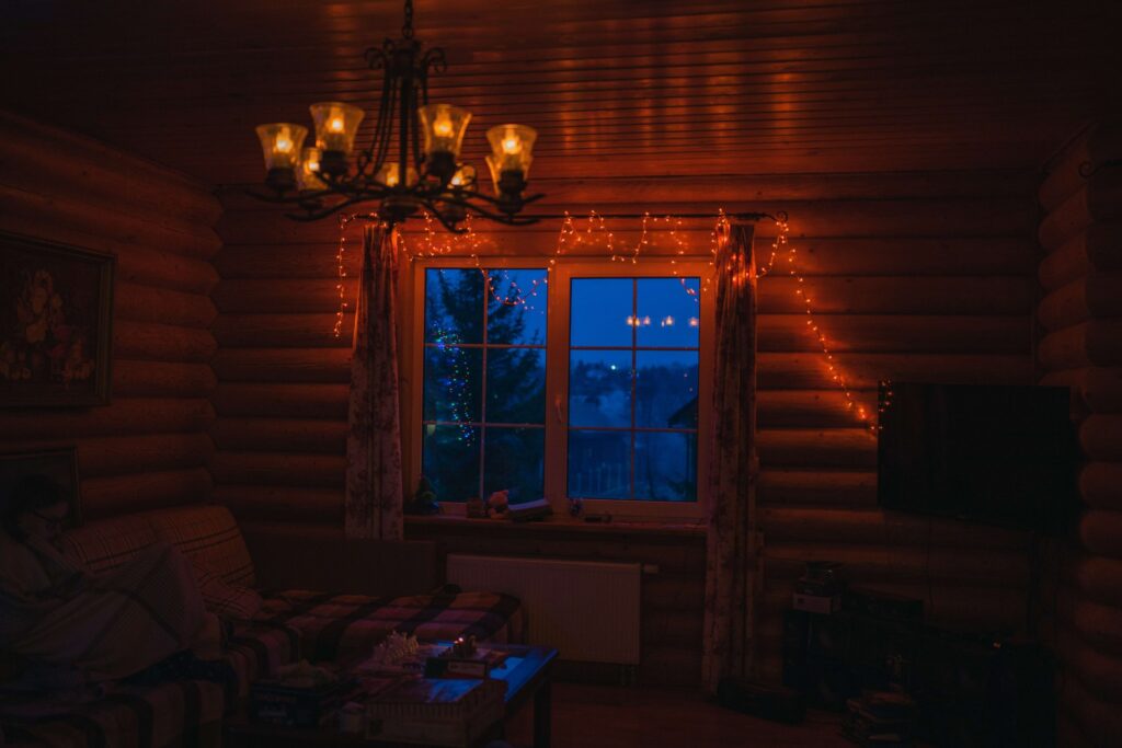 A cozy room with wooden walls is softly lit by a chandelier, evoking the charm of Thanksgiving. An evening view through the window, adorned with string lights, enhances the scene like a cheerful cartoon. A couch and a table with items are in the foreground, creating a warm, inviting atmosphere.