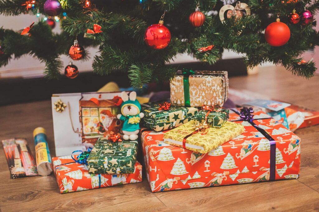 A variety of colorful screen-free gifts are nestled under a decorated Christmas tree. The tree is adorned with red, gold, and orange ornaments. A plush bunny toy is visible among the presents, adding to the festive scene.