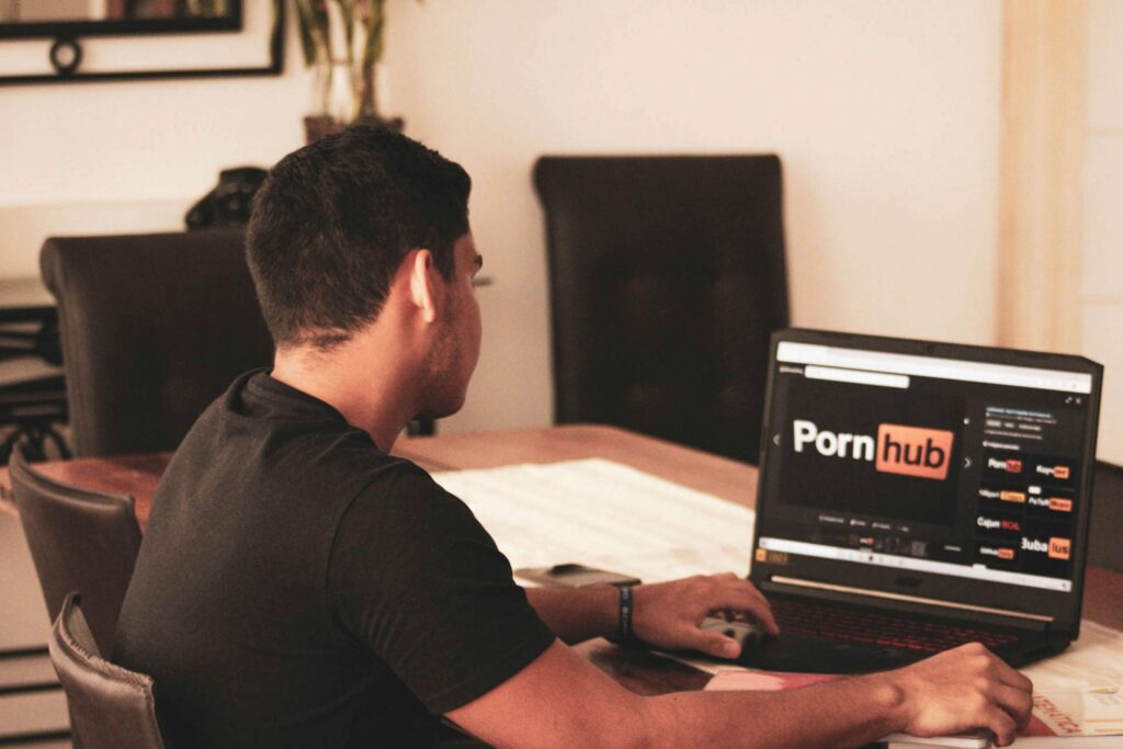 A person is seated at a table, using a laptop that displays an adult content website's homepage. Despite the room's minimal decor and neutral palette, the focus on No Porn November adds a layer of contemplation to the setting, urging mindful digital interactions.