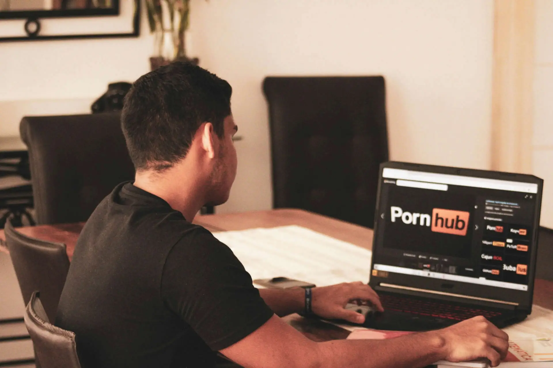 A person is seated at a table, using a laptop that displays an adult content website's homepage. Despite the room's minimal decor and neutral palette, the focus on No Porn November adds a layer of contemplation to the setting, urging mindful digital interactions.