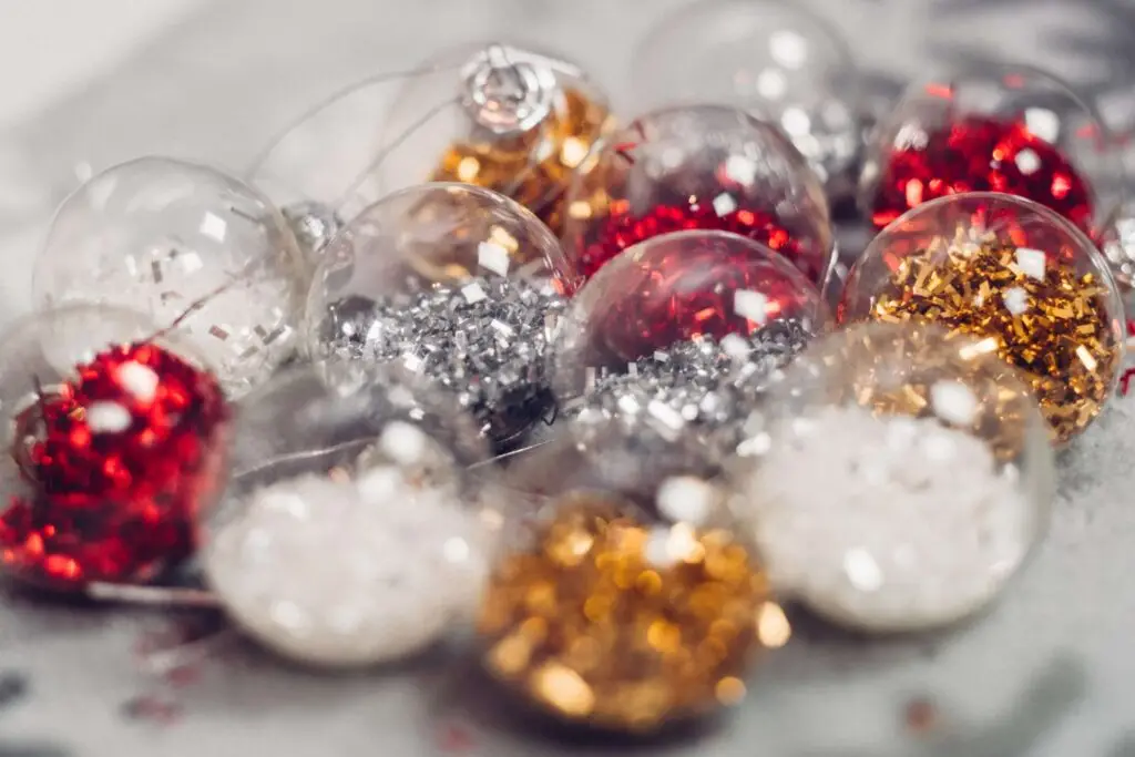 Close-up of small, transparent Christmas ornaments filled with festive glitter in red, silver, and gold. Scattered like a creative Christmas charcuterie board on a gray surface, they create a sparkling and colorful holiday display.