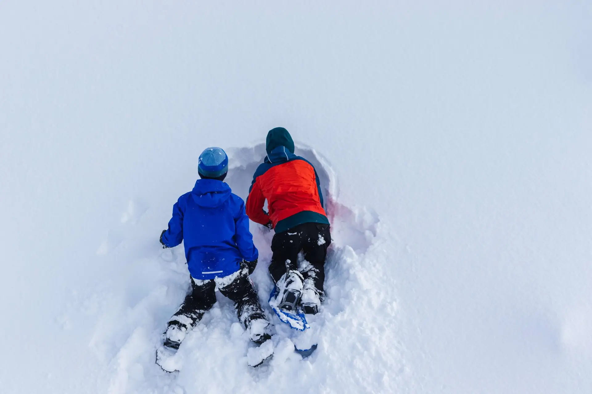 Two children are lying in the snow, snug in winter jackets, snow boots, and cozy winter hats for kids. They're digging into a snowbank, one in a blue jacket and the other in red. The surrounding area is blanketed with fresh, undisturbed snow.