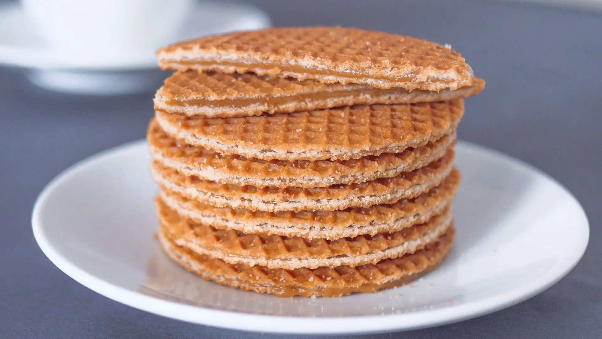 A stack of stroopwafels sits on a white plate, reminiscent of a festive Christmas Crack Recipe, with a cup blurred in the background. The waffles are layered with caramel syrup, arranged neatly one on top of the other on a dark surface.