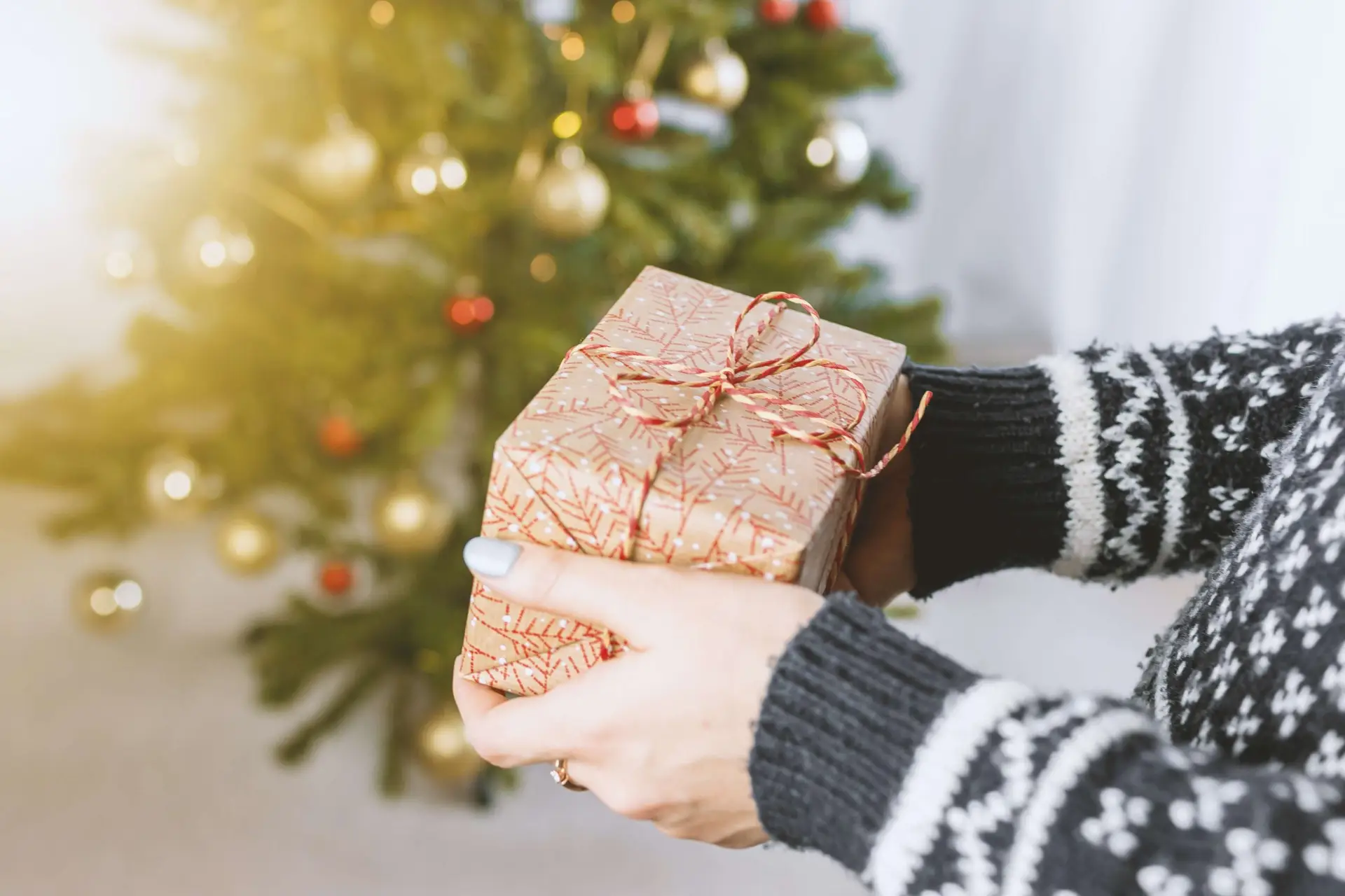 A person in a festive sweater holds a screen-free gift wrapped with a red ribbon, standing before a decorated Christmas tree adorned with gold and red ornaments.