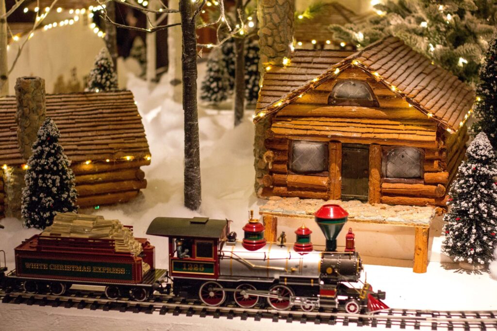 A festive miniature scene showcases a toy train labeled White Christmas Express on snowy tracks. Nearby, log cabins are decked with twinkling lights, surrounded by small snow-dusted pine trees, perfect for inspiring Christmas party activities for kids.