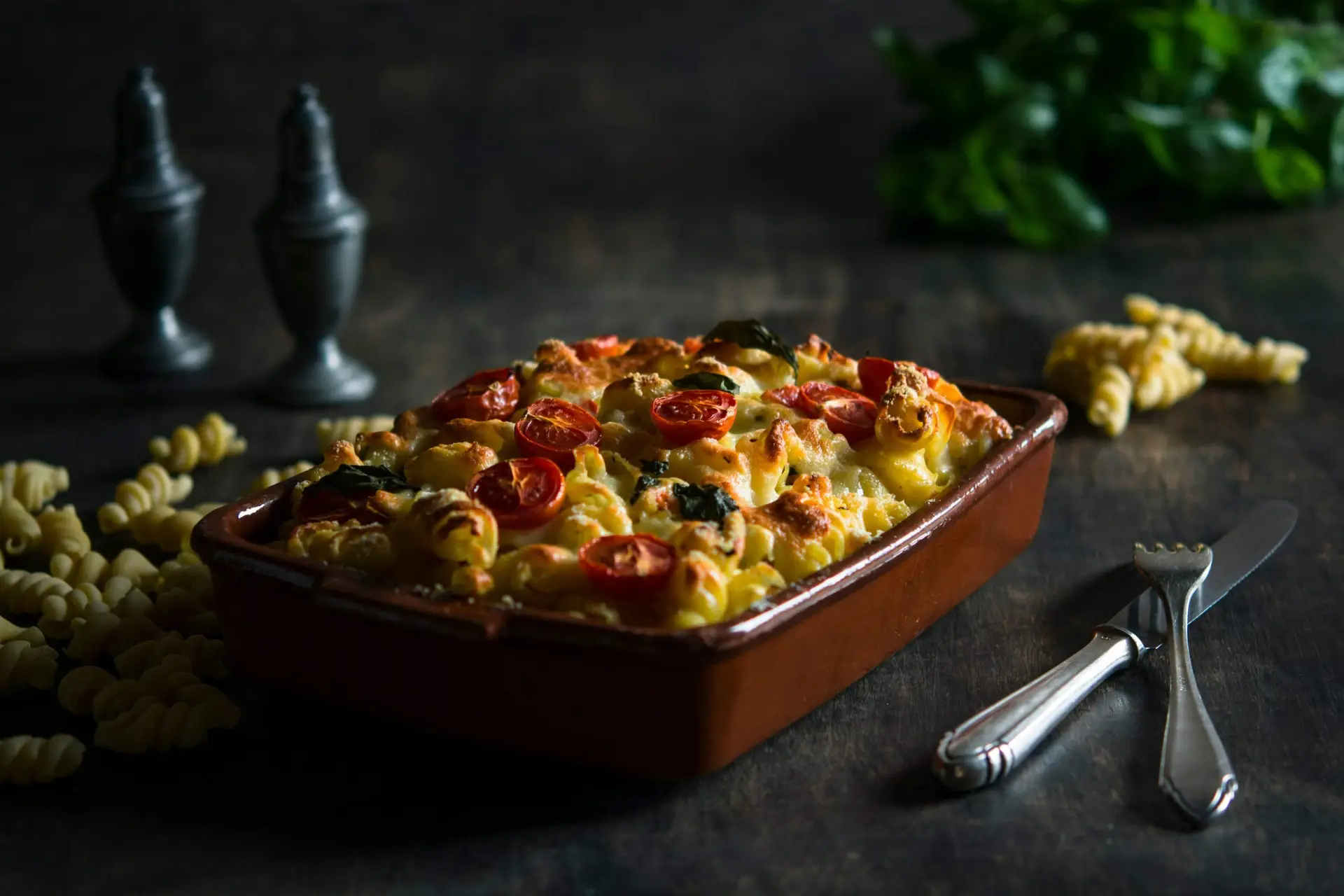 A baked pasta dish with melted cheese and cherry tomatoes rests in a rectangular dish, embodying the warmth of Christmas casserole recipes. The table also holds uncooked pasta spirals, silverware, and salt and pepper shakers, with a bunch of fresh greens in the background.