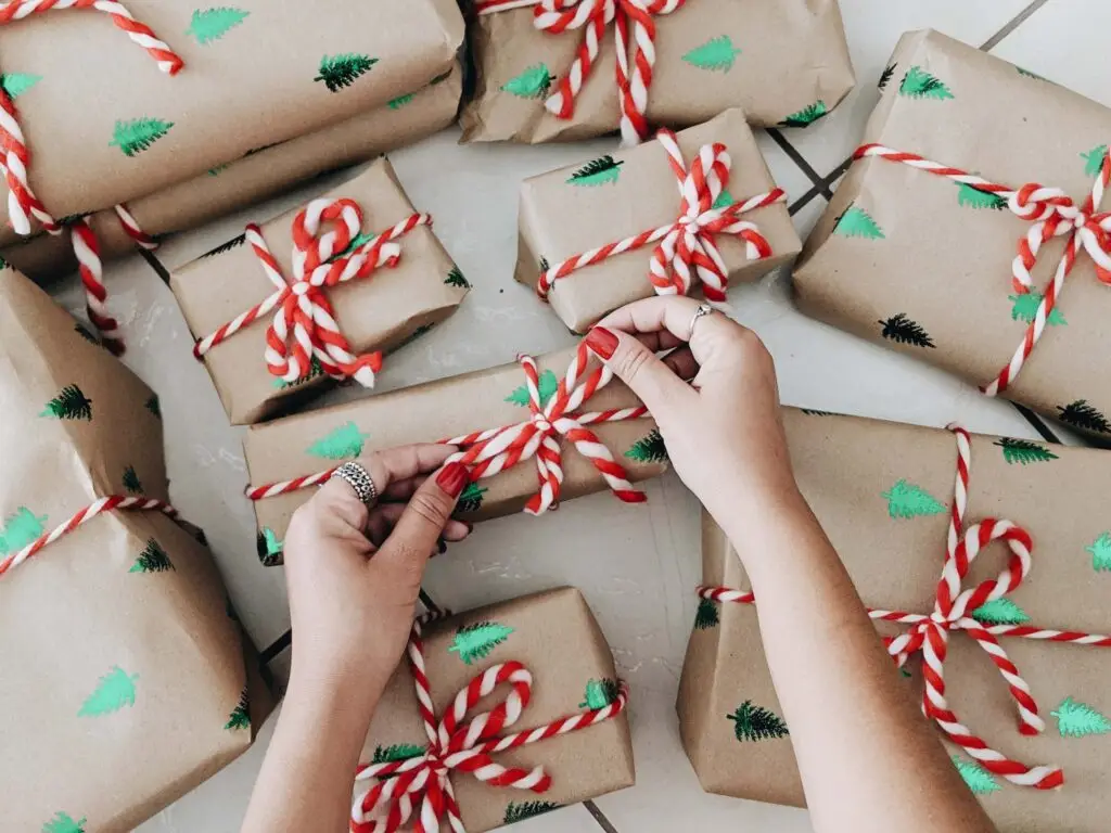 A person ties a red and white striped ribbon onto a brown gift wrapped in paper adorned with green Christmas tree prints. Several similar screen-free gifts are arranged around, all set against a light-colored floor for that perfect festive touch.