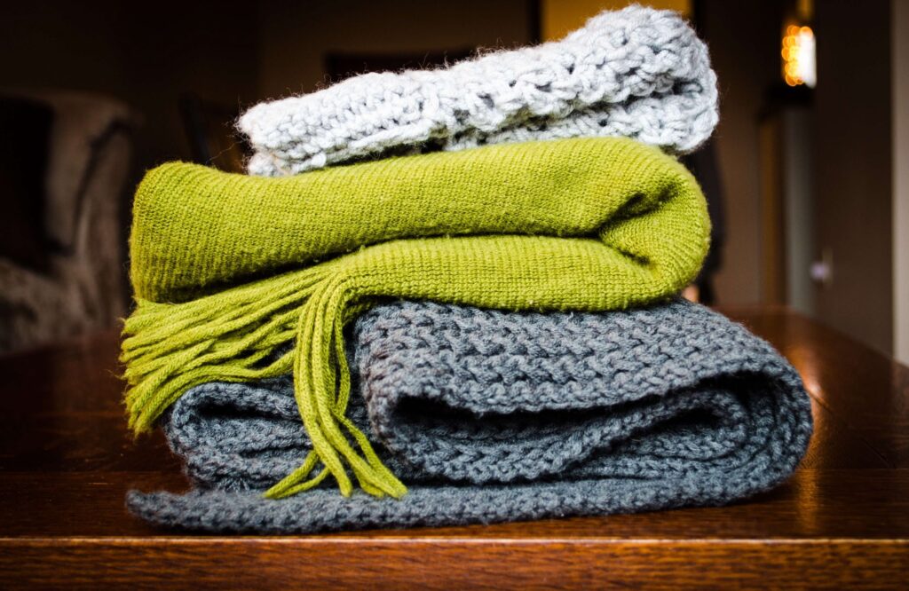 A stack of three folded knit blankets, reminiscent of cozy kids' winter gear, sits on a wooden table. The pile features a white blanket on top, a green fringed one in the middle, and a dark gray blanket at the bottom against a softly blurred room backdrop.