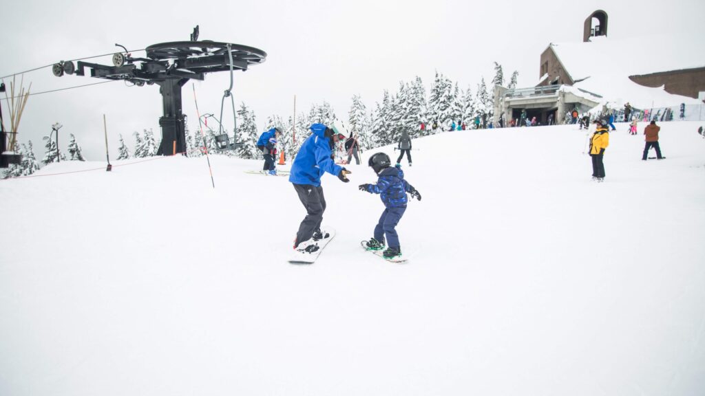 Two people joyfully snowboard together on a snowy slope near a ski lift, showcasing the thrill of winter sports for kids. The larger person in a blue jacket assists the smaller one in dark clothing. Snow-covered trees and a mountain lodge decorate the background as others enjoy the area.