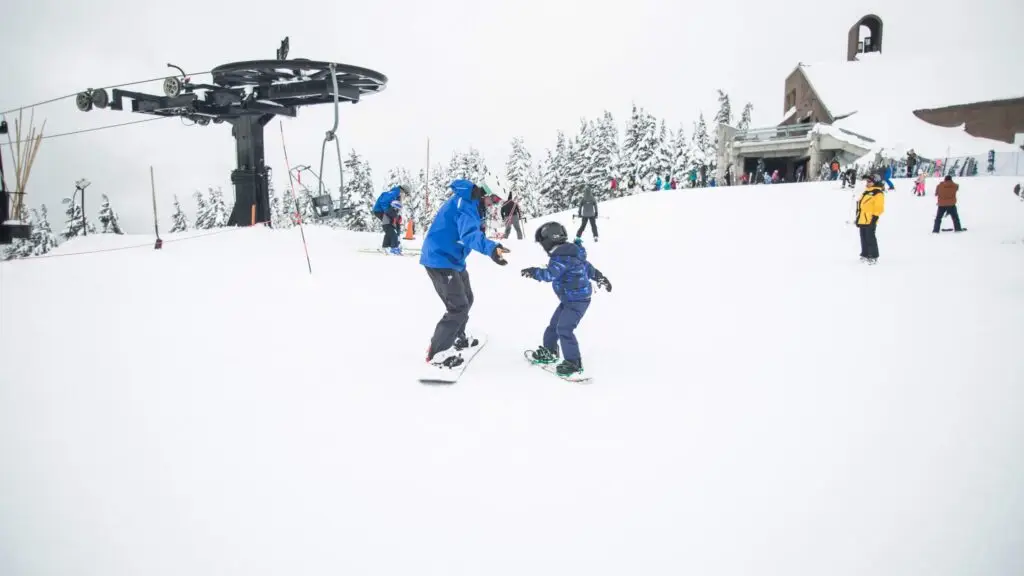 Two people joyfully snowboard together on a snowy slope near a ski lift, showcasing the thrill of winter sports for kids. The larger person in a blue jacket assists the smaller one in dark clothing. Snow-covered trees and a mountain lodge decorate the background as others enjoy the area.