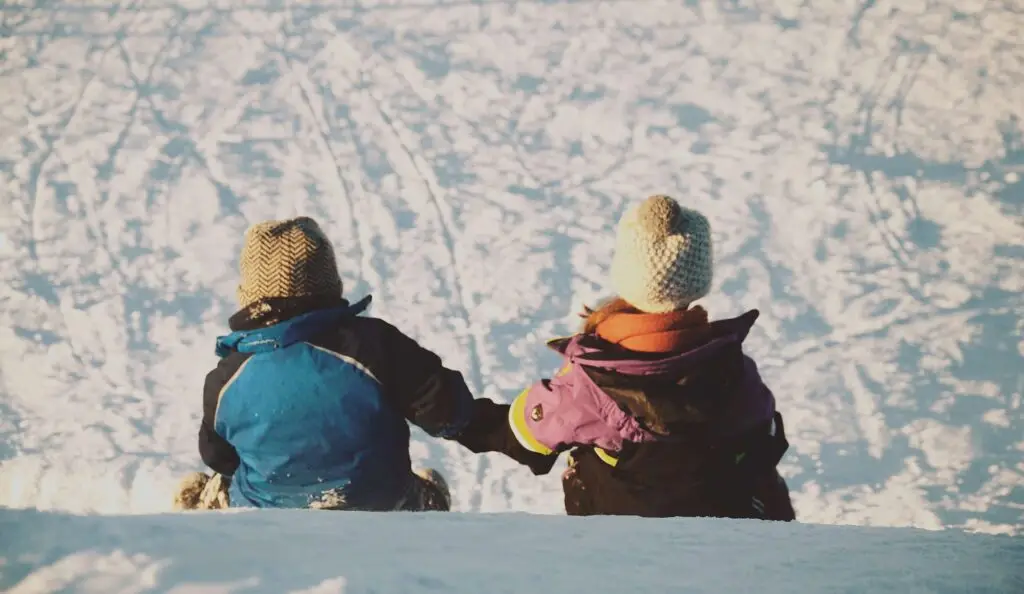 Two children in winter clothing and knitted hats sit on a snowy slope, holding hands. They are facing away from the camera, perhaps sharing winter jokes for kids as they gaze at the snowy landscape ahead.