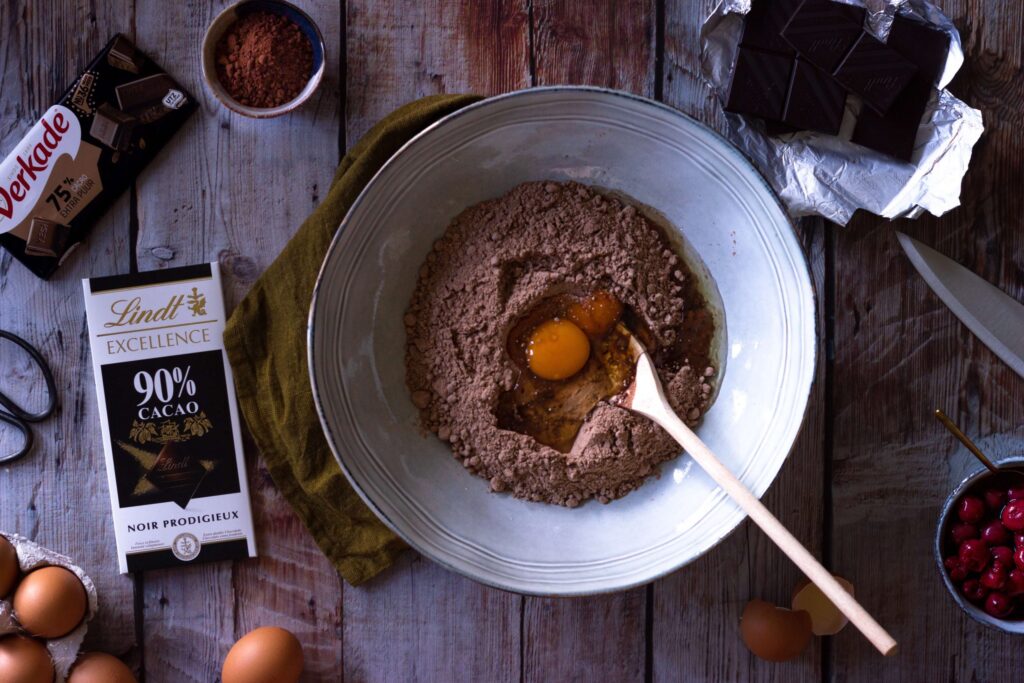 A bowl with cocoa powder and cracked eggs ready for mixing sits on a wooden surface, surrounded by dark chocolate, a knife, eggshells, and cocoa powder—perfect for crafting a delectable Christmas tree snack cake. An open chocolate bar wrapper and a green cloth are nearby.